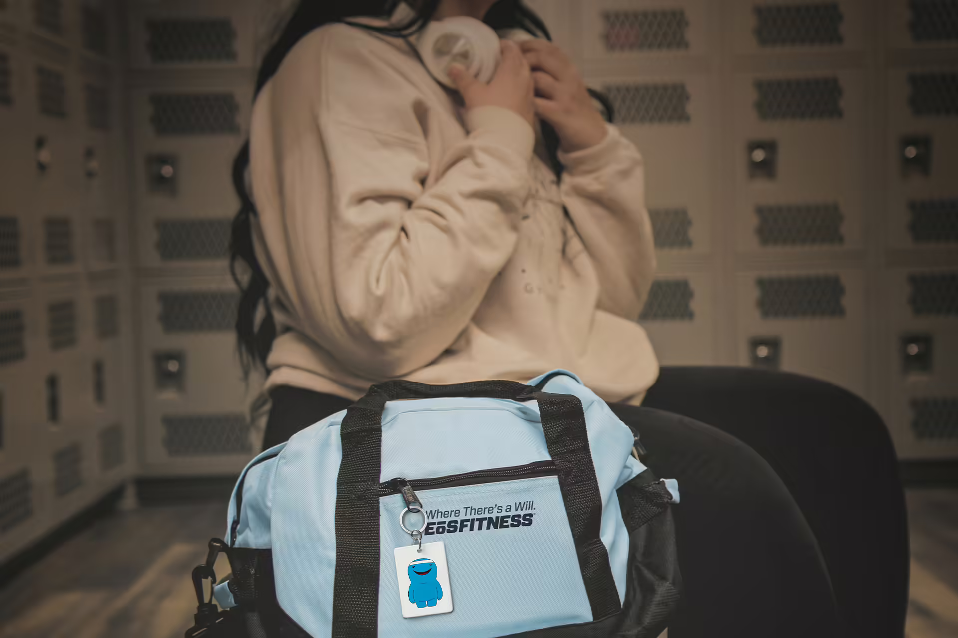 A woman sitting on a bench in a gym locker room with an EOS duffle bag next to her.