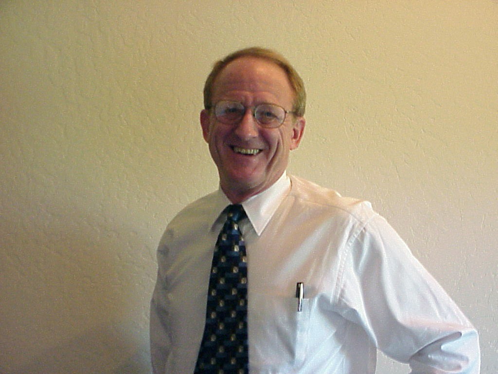 A photo of a man smiling and dressed in a fine white dress shirt with a blue tie and a pen in his front left pocket.